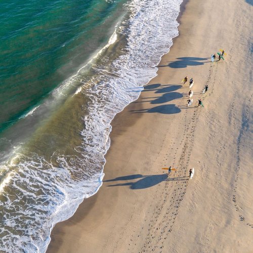 Aerial view of people walking and surfing on a sandy beach with waves crashing ashore. outer-banks-lifestyle